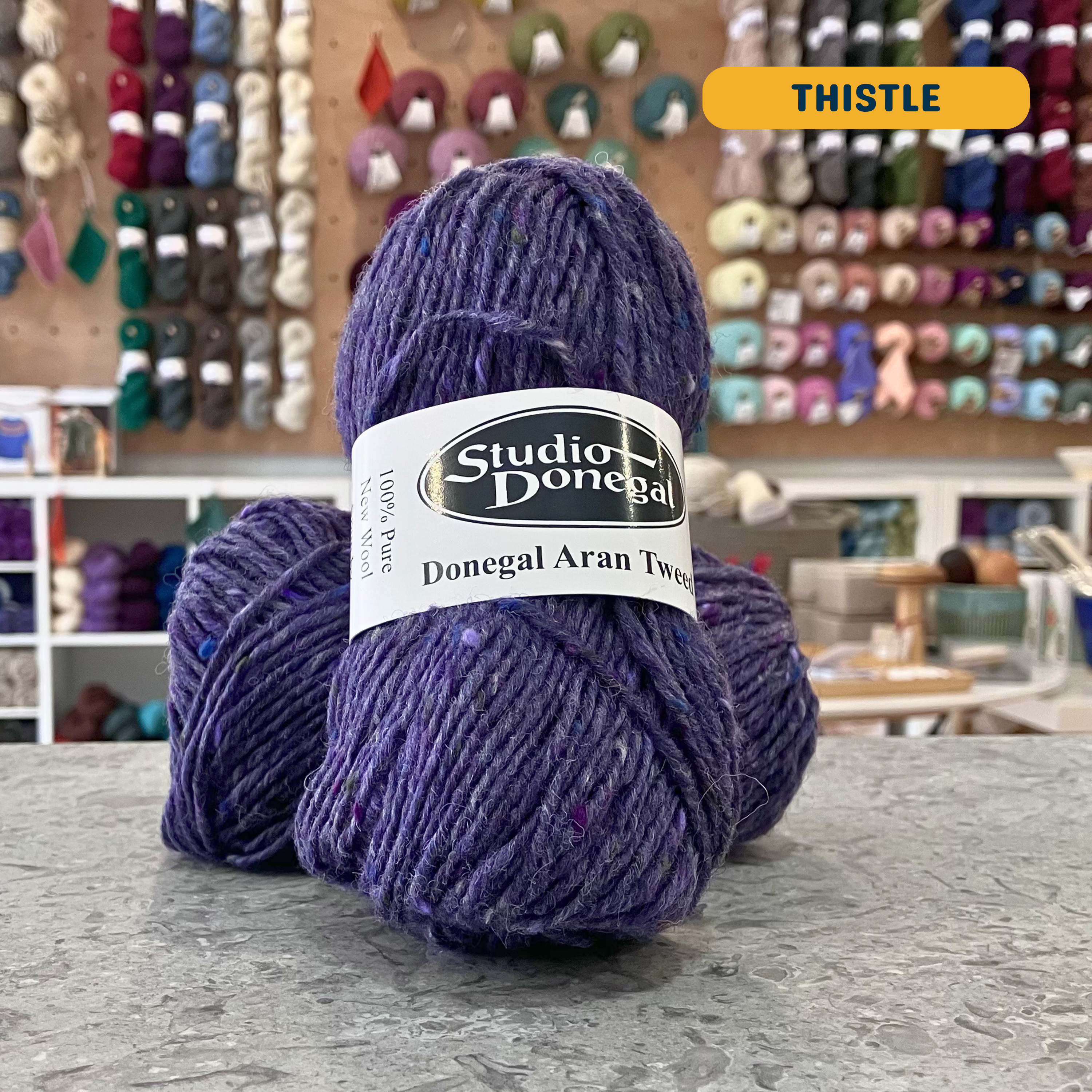 50g purple-grey (thistle) with multi-coloured specks Irish wool aran yarn, 'Studio Donegal Donegal Aran Tweed' label visible, craft shop background (blurred)