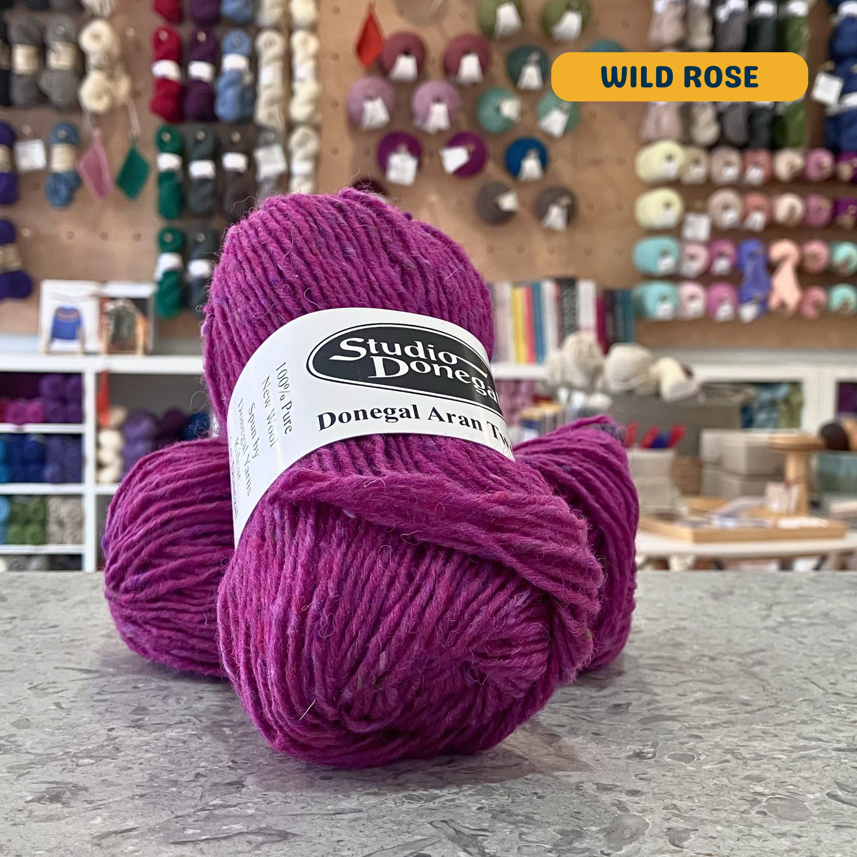 50g pink (wild rose) with multi-coloured specks Irish wool aran yarn, 'Studio Donegal Donegal Aran Tweed' label visible, craft shop background (blurred)