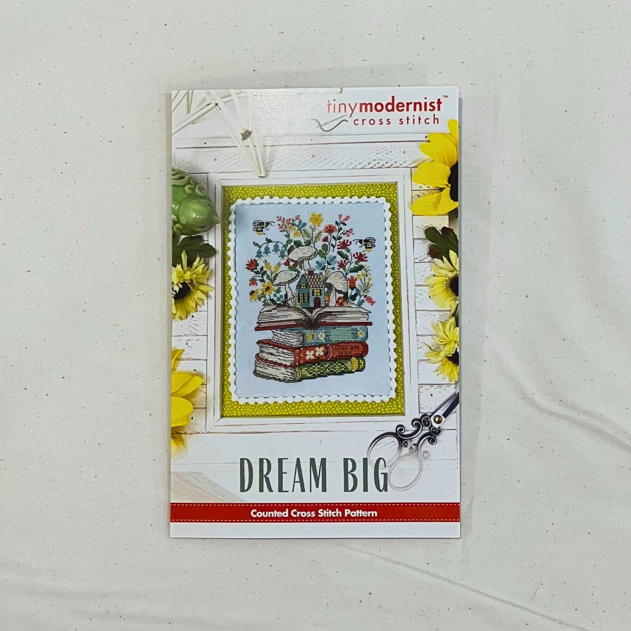 A counted cross stitch pattern card for 'Dream Big' by Tiny Modernist Cross Stitch, featuring a finished sample with central stitched design of birds on stacked books with flowers.