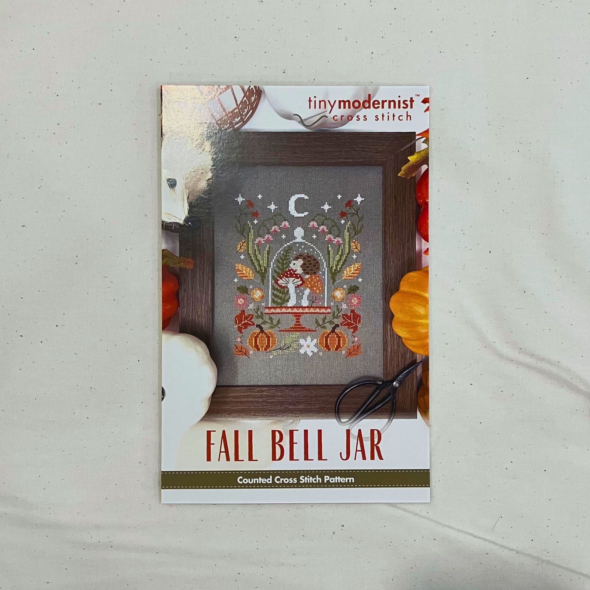 A counted cross stitch pattern card for 'Fall Bell Jar' by Tiny Modernist Cross Stitch, featuring a finished sample of a hedgehog, mushrooms, pumpkins, and more.