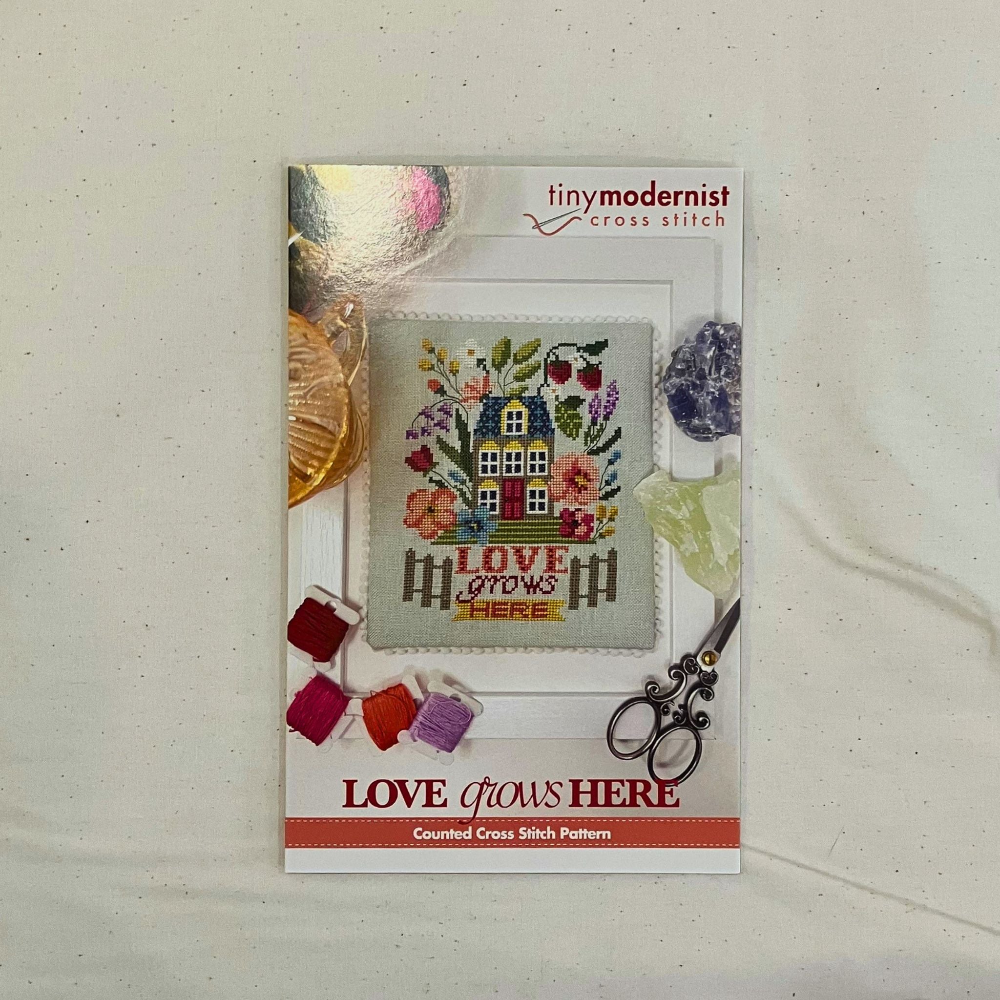 A counted cross stitch pattern card for 'Love Grows Here' by Tiny Modernist Cross Stitch, featuring a finished sample of  a colorful embroidered house and floral design.