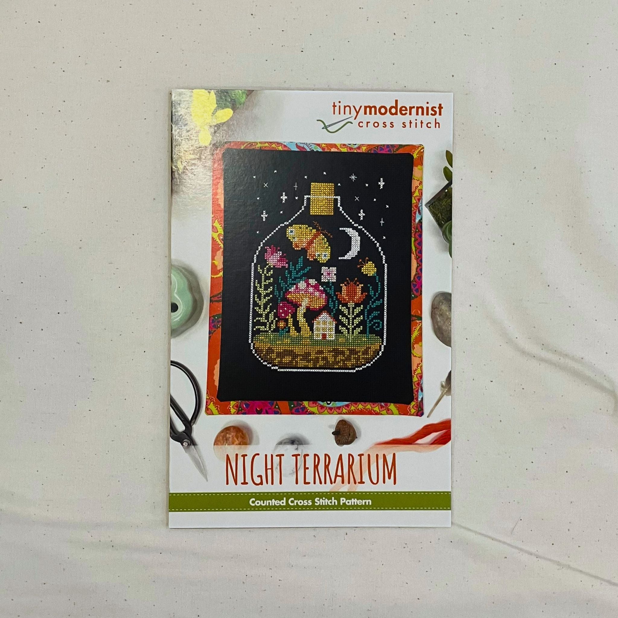 A counted cross stitch pattern card for 'Night Terrarium' by Tiny Modernist Cross Stitch, featuring a finished sample of a colourful illustration of a mushroom garden inside a glass bottle under a night sky.