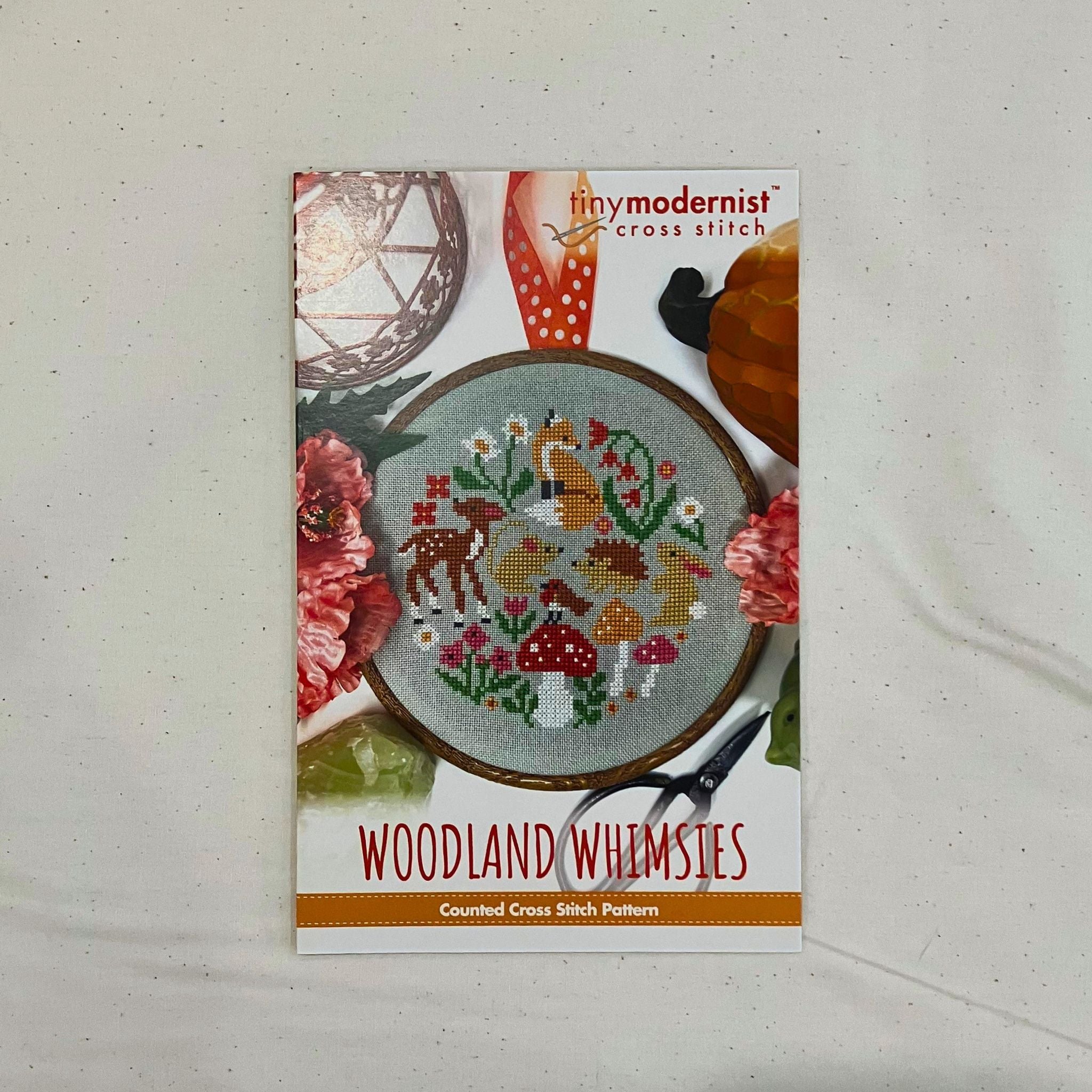 A counted cross stitch pattern card for 'Woodland Whimsies' by Tiny Modernist Cross Stitch, featuring a finished sample of a colourful cross-stitched woodland design with deer, mushrooms, flowers, and a fox.