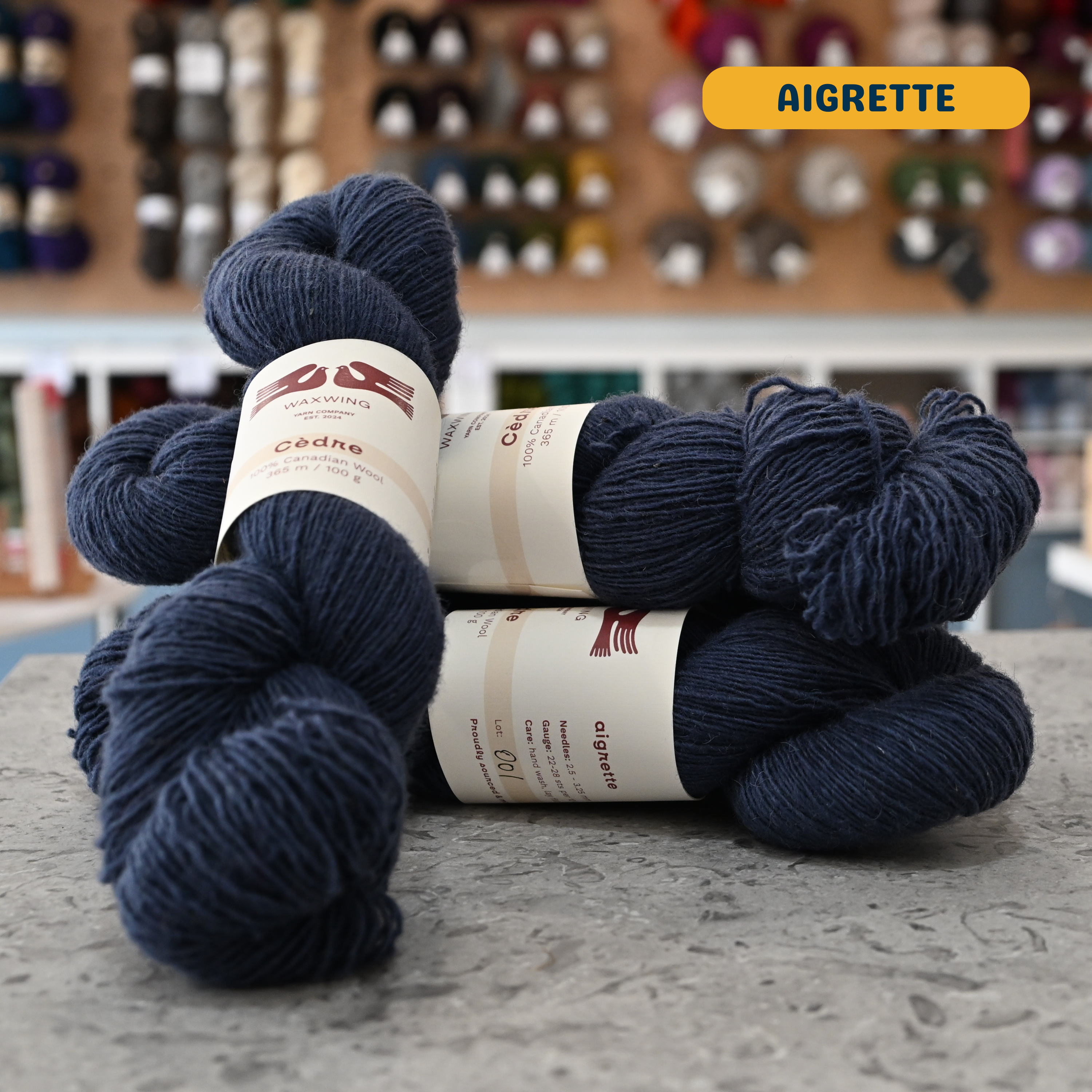 100g dark blue (aigrette) canadian wool fingering yarn, 'Waxwing Yarn Company Cèdre' label visible, craft shop background (blurred)