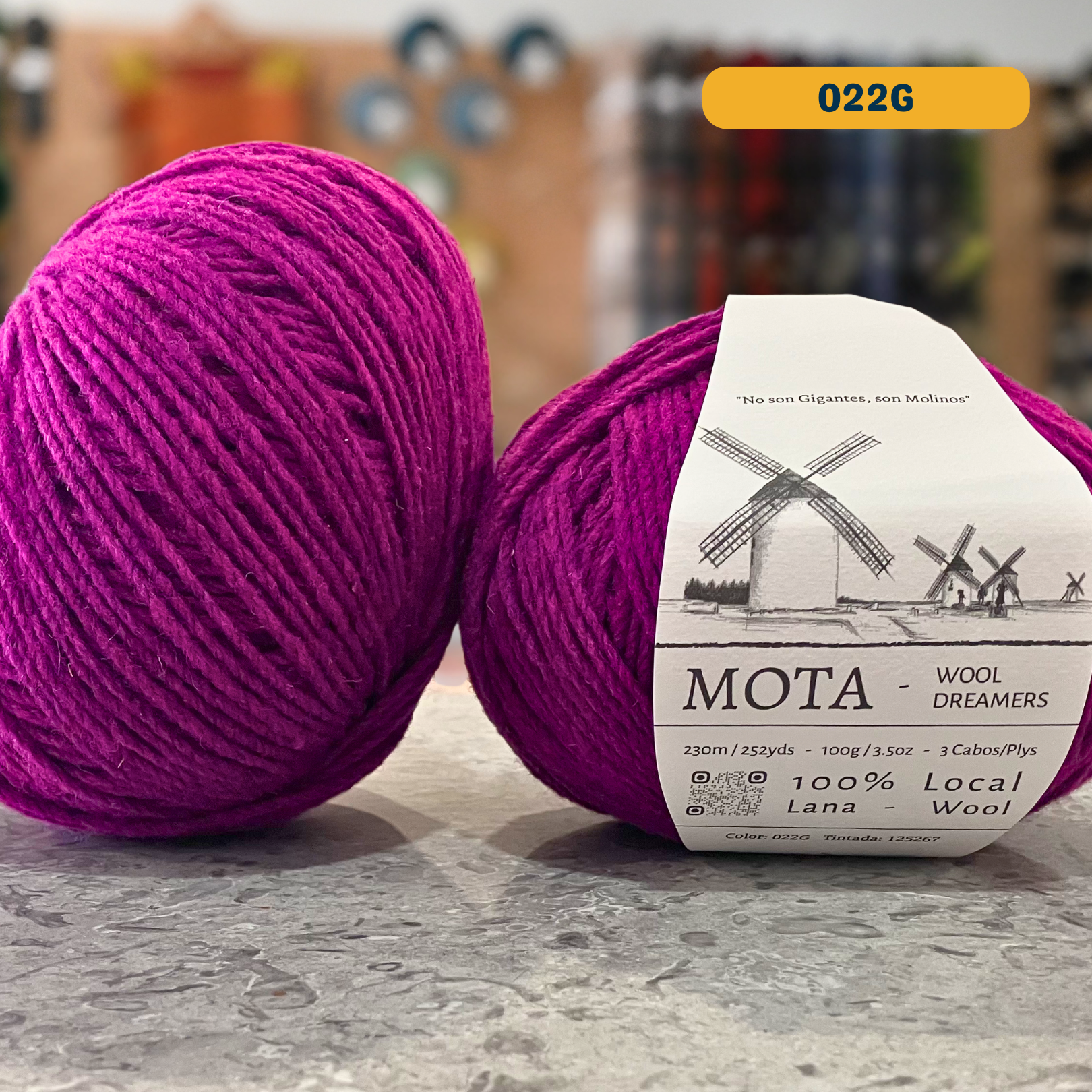 100g vibrant pink-purple (022G) merino & manchega wool yarn, 'Mota Wooldreamers 100% Wool' label visible, craft shop background (blurred)
