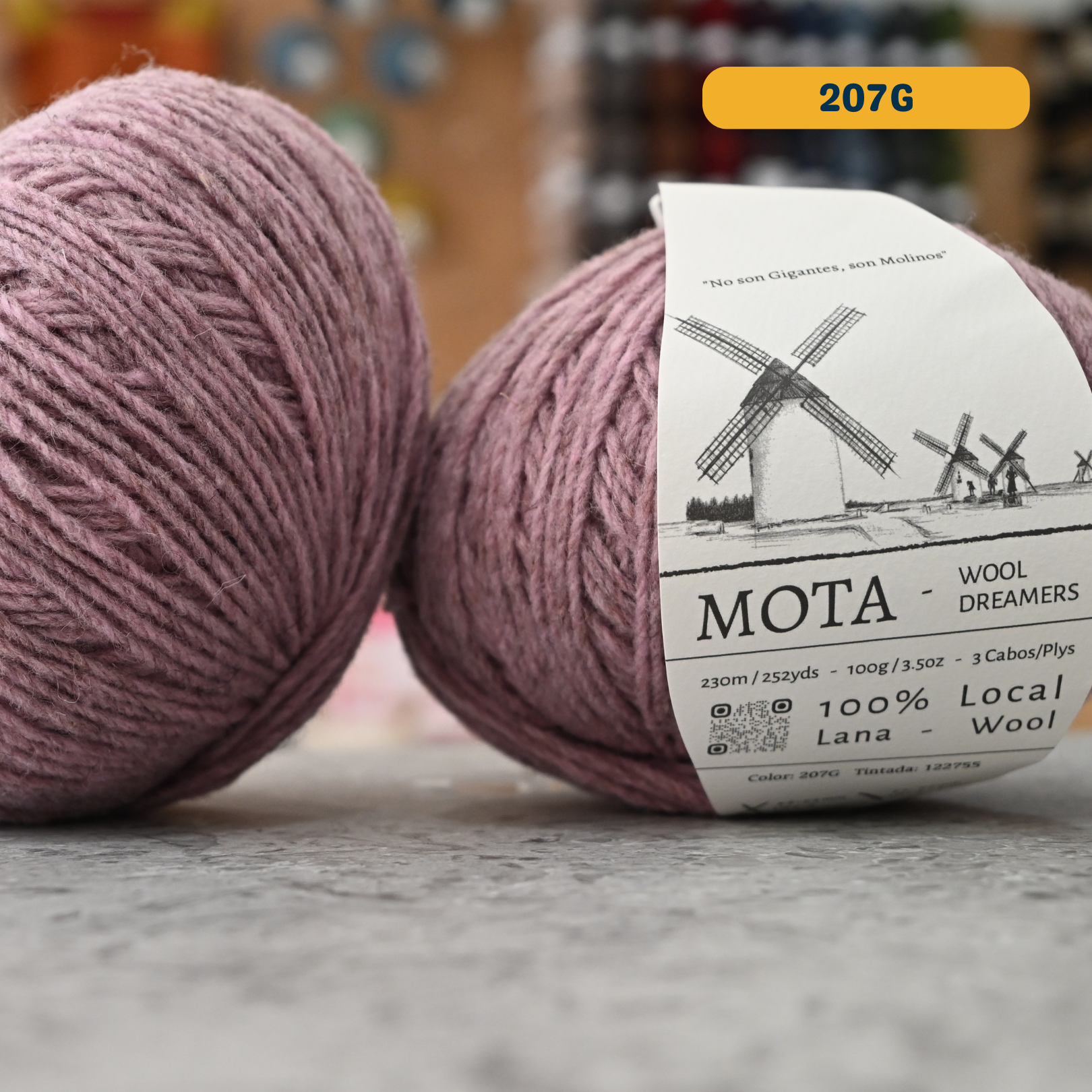 100g grey-pink (207G)  merino & manchega wool yarn, 'Mota Wooldreamers 100% Wool' label visible, craft shop background (blurred)
