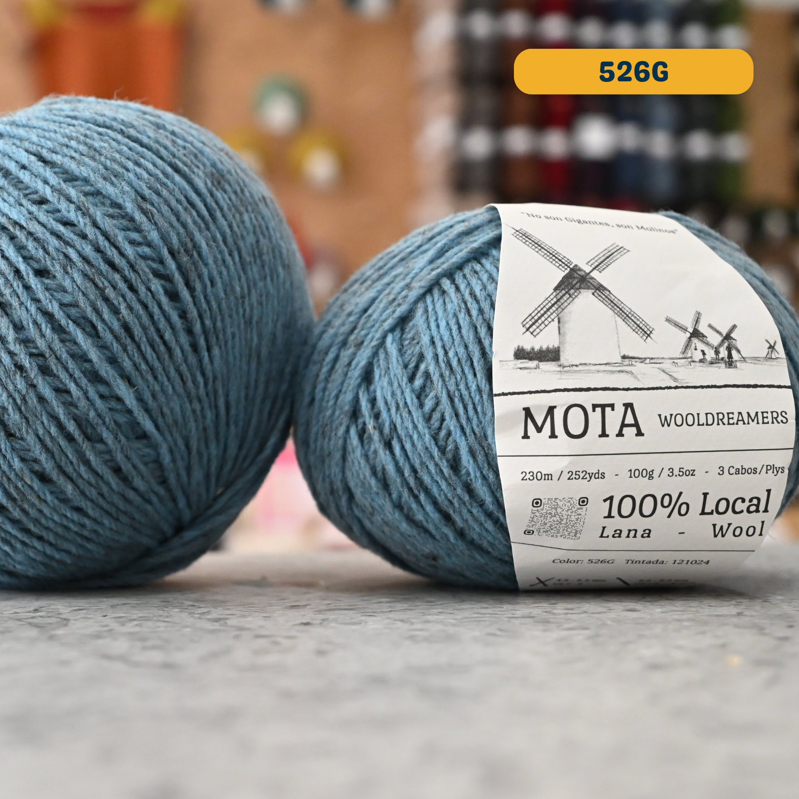 100g blue-grey (526G) merino & manchega wool yarn, 'Mota Wooldreamers 100% Wool' label visible, craft shop background (blurred)

