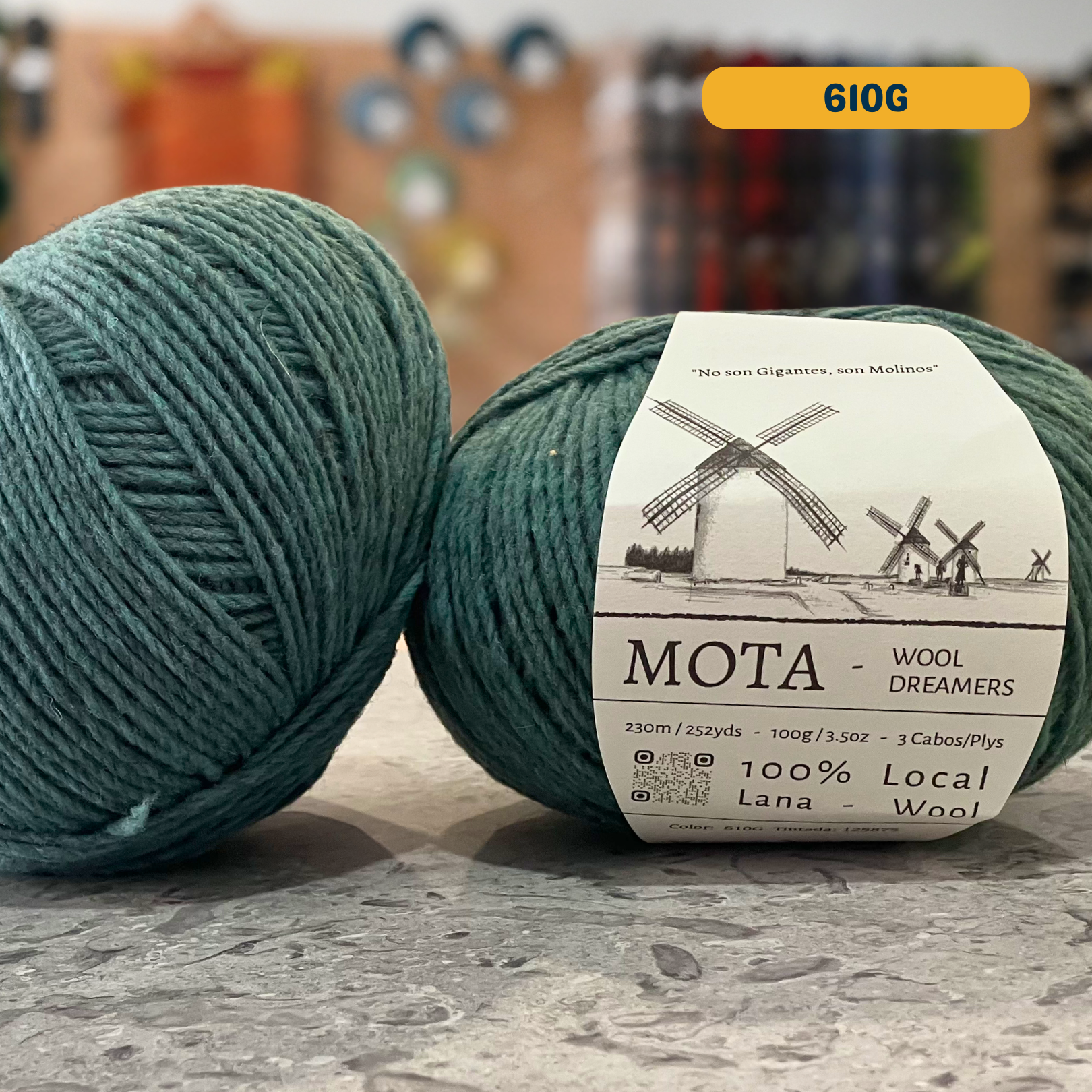 100g grey-teal (610G) merino & manchega wool yarn, 'Mota Wooldreamers 100% Wool' label visible, craft shop background (blurred)
