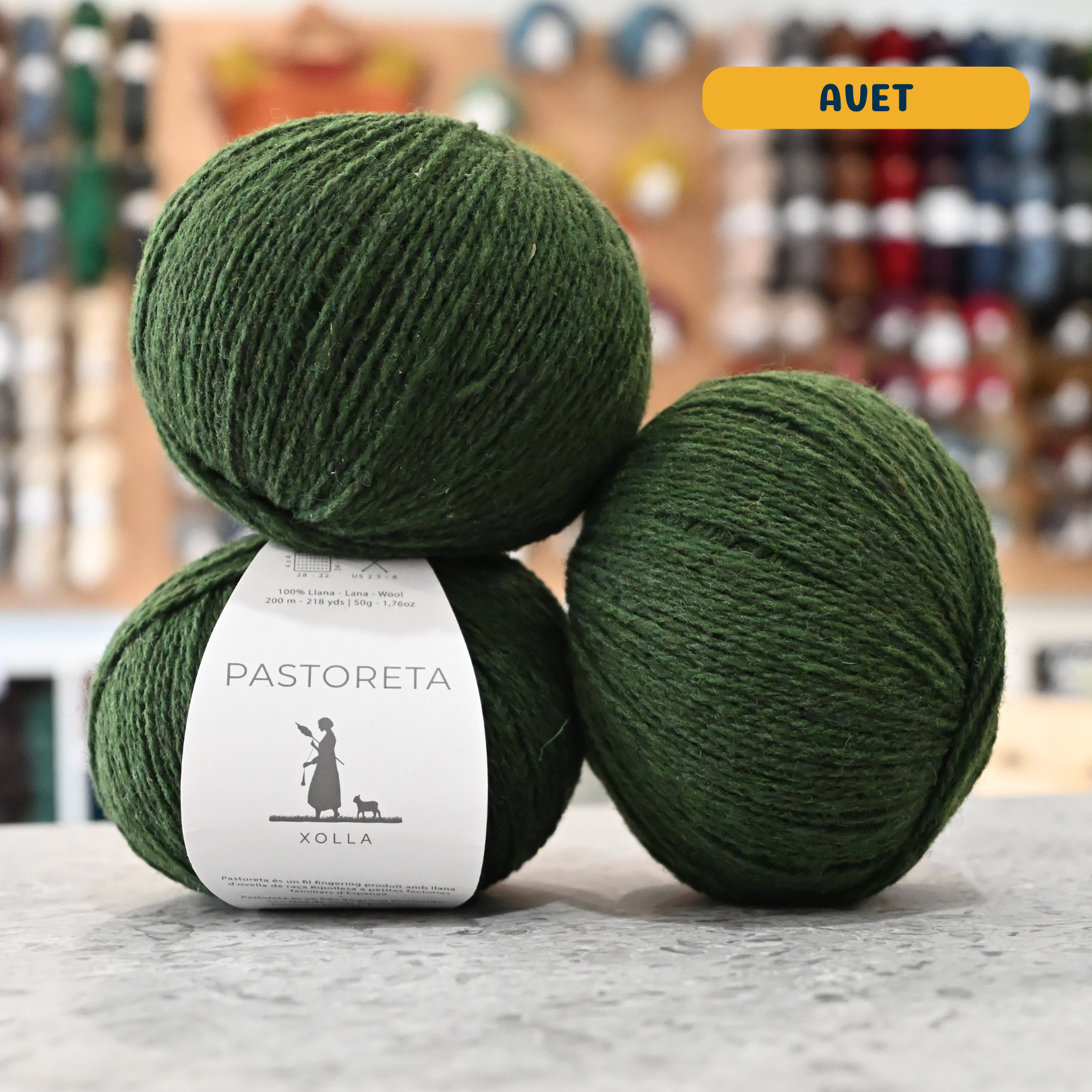 50g green (avet) Spanish wool fingering yarn, 'Pastoreta Xolla' label visible, craft shop background (blurred)