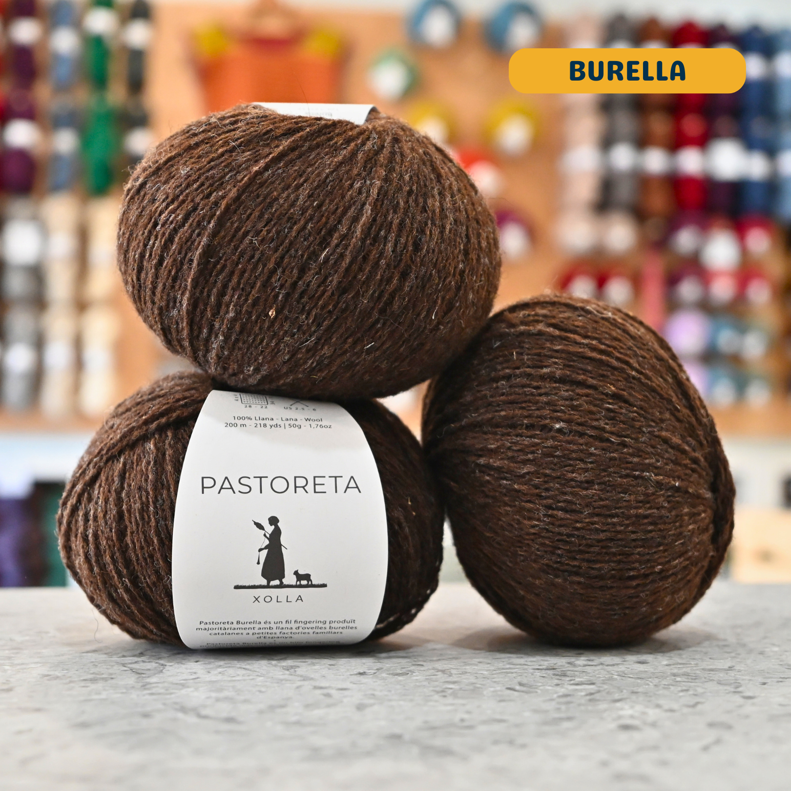 50g caramel-brown (burella) Spanish wool fingering yarn, 'Pastoreta Xolla' label visible, craft shop background (blurred)

