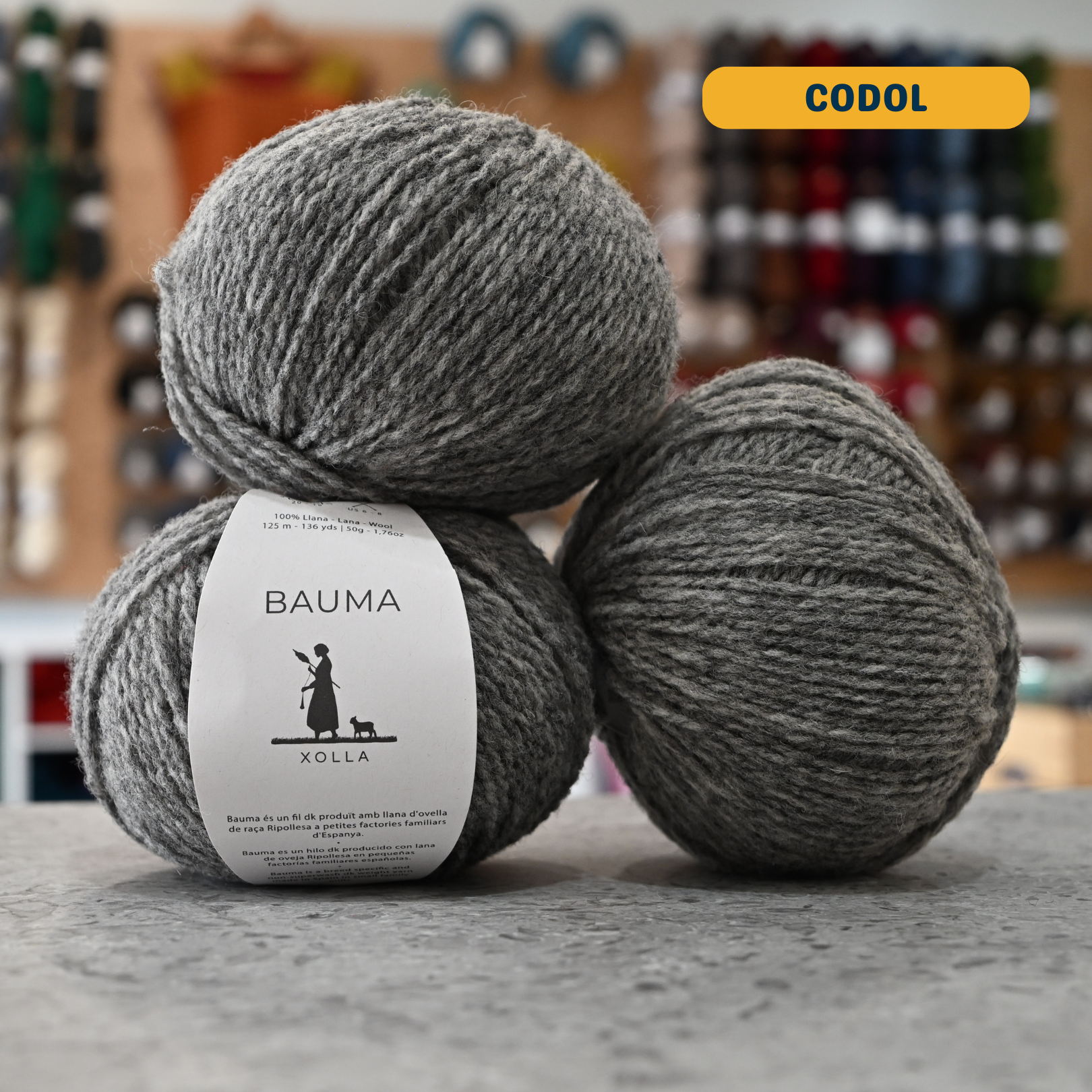 50g grey (codol) Spanish wool DK yarn, 'Bauma Xolla' label visible, craft shop background (blurred)