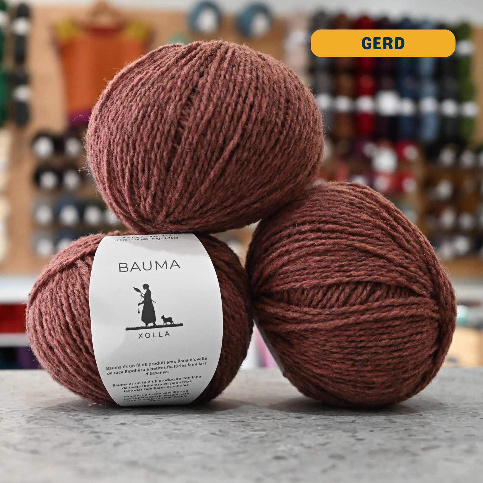 50g grey-pink (gerd) Spanish wool DK yarn, 'Bauma Xolla' label visible, craft shop background (blurred)