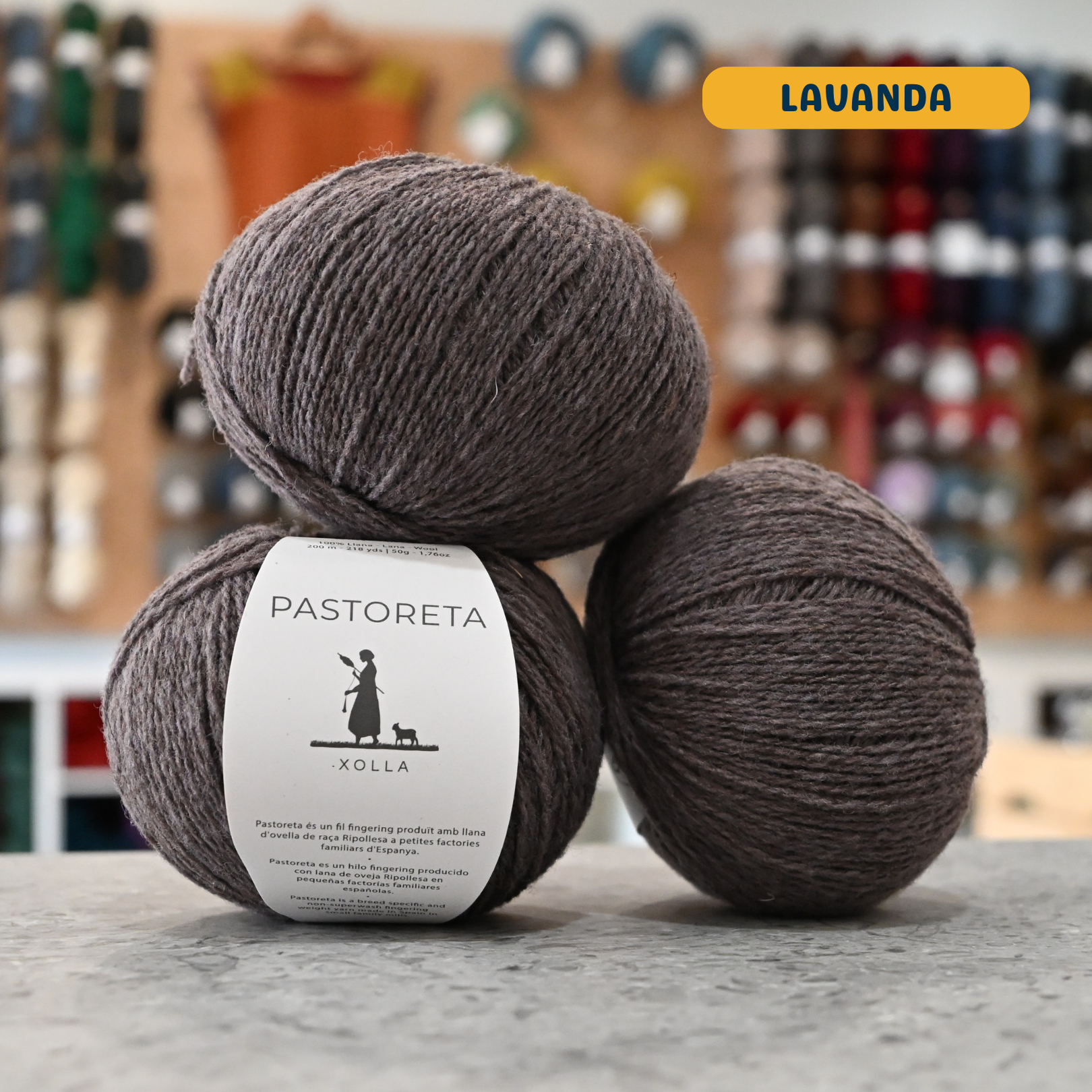50g purple-grey (lavanda) Spanish wool fingering yarn, 'Pastoreta Xolla' label visible, craft shop background (blurred)
