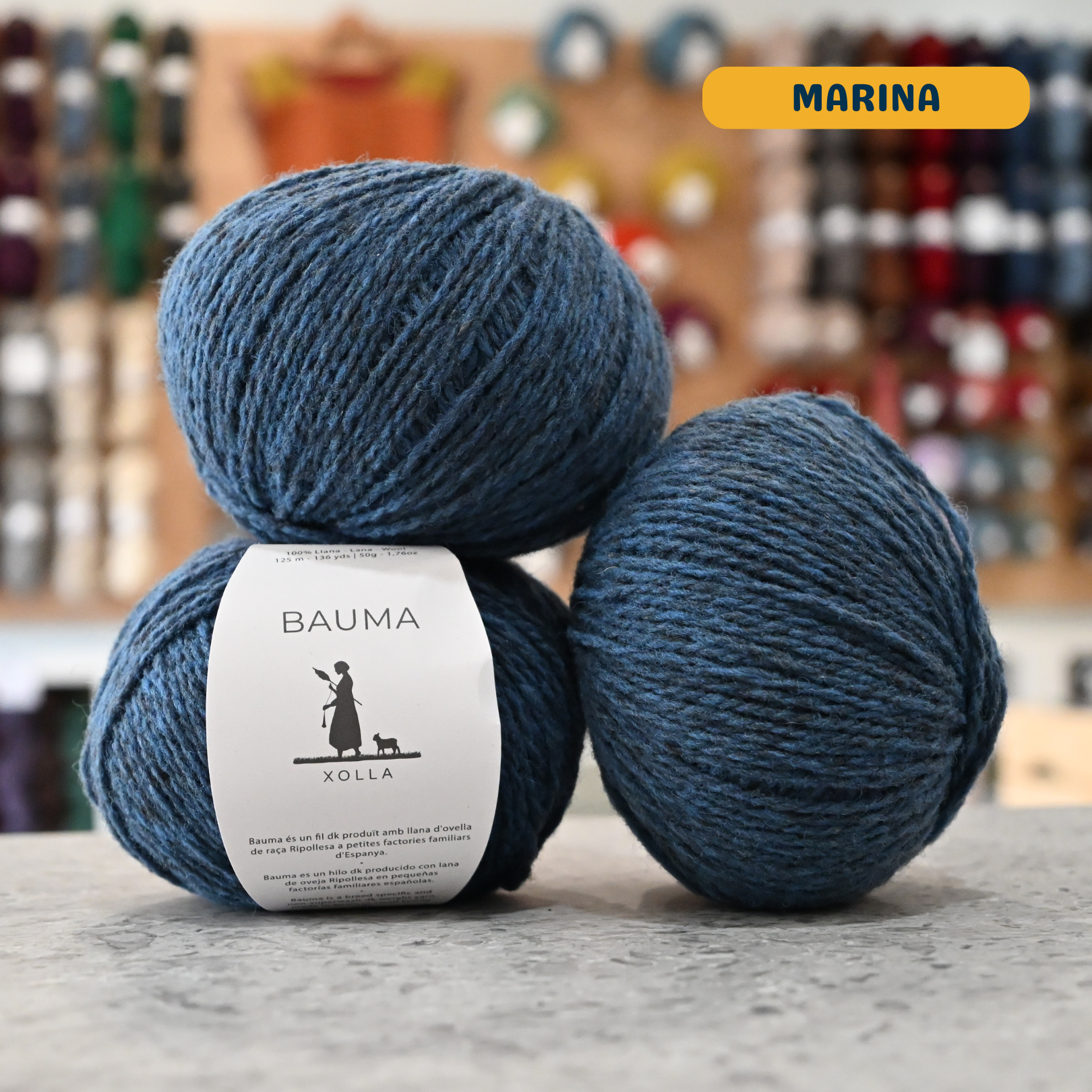 50g blue (marina) Spanish wool DK yarn, 'Bauma Xolla' label visible, craft shop background (blurred)