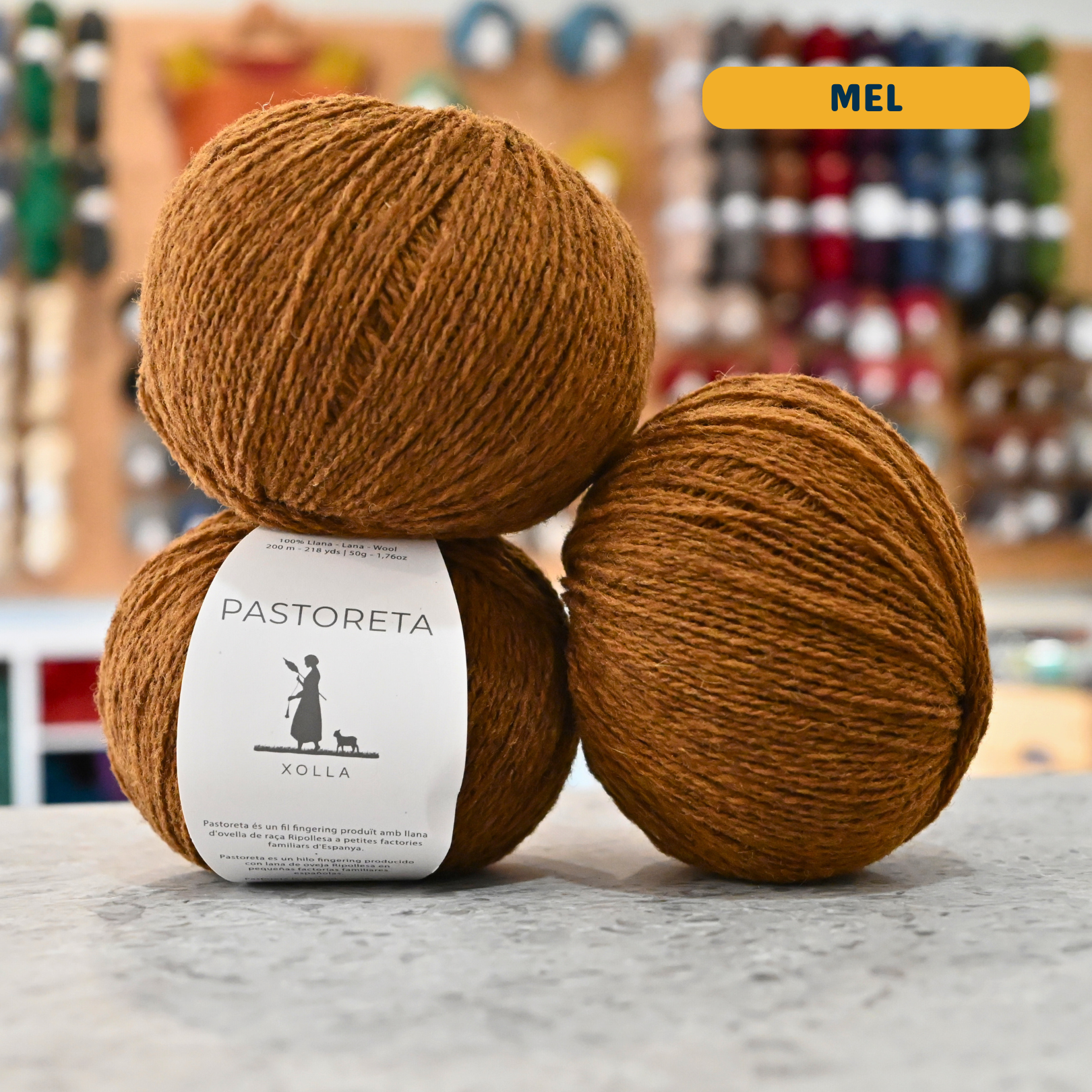50g orange-brown (mel) Spanish wool fingering yarn, 'Pastoreta Xolla' label visible, craft shop background (blurred)
