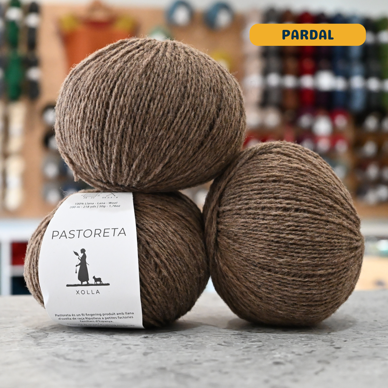 50g brown-beige (pardal) Spanish wool fingering yarn, 'Pastoreta Xolla' label visible, craft shop background (blurred)
