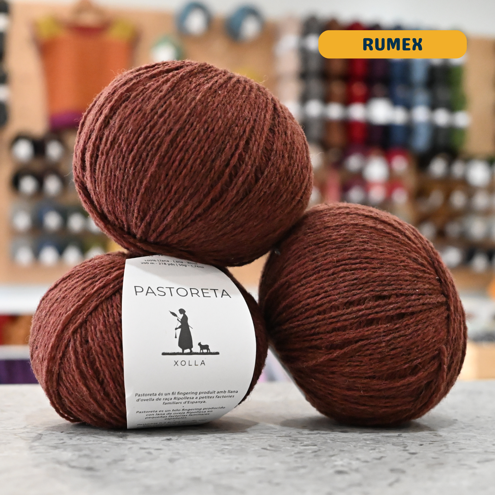 50g brown-red (rumex) Spanish wool fingering yarn, 'Pastoreta Xolla' label visible, craft shop background (blurred)
