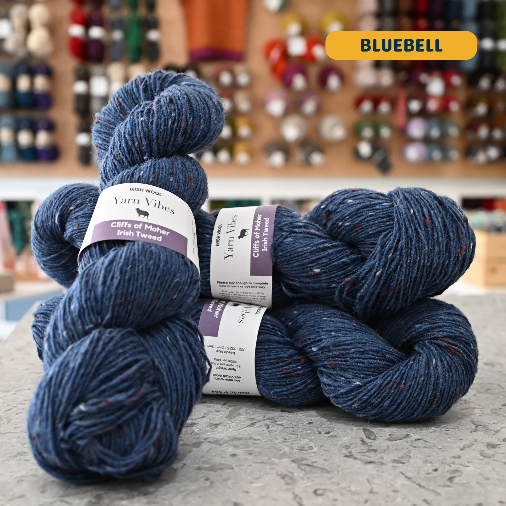 100g dark blue with rainbow specks (bluebell) Irish wool merino sport yarn, 'Yarn Vibes Cliffs of Moher Irish Tweed' label visible, craft shop background (blurred)
