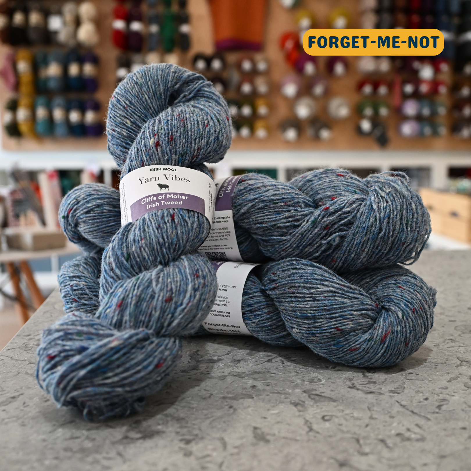 100g light blue with rainbow specks (forget-me-not) Irish wool merino sport yarn, 'Yarn Vibes Cliffs of Moher Irish Tweed' label visible, craft shop background (blurred)
