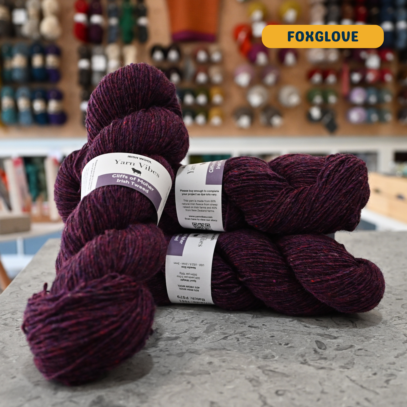 100g dark purple (foxglove) Irish wool merino sport yarn, 'Yarn Vibes Cliffs of Moher Irish Tweed' label visible, craft shop background (blurred)