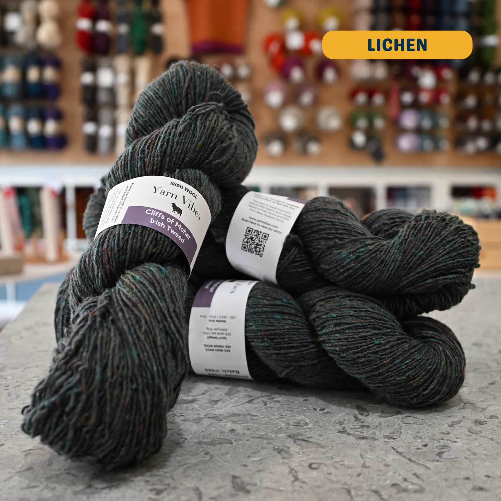 100g grey-green (lichen) Irish wool merino sport yarn, 'Yarn Vibes Cliffs of Moher Irish Tweed' label visible, craft shop background (blurred)
