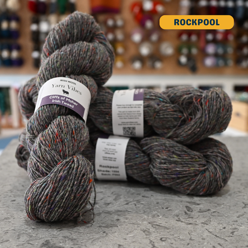 100g grey with rainbow specks (rockpool) Irish wool merino sport yarn, 'Yarn Vibes Cliffs of Moher Irish Tweed' label visible, craft shop background (blurred)