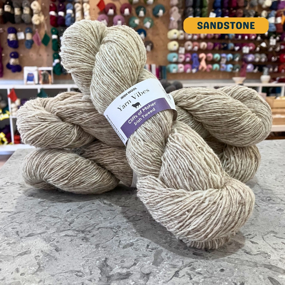 100g heathered beige (sandstone) Irish wool merino sport yarn, 'Yarn Vibes Cliffs of Moher Irish Tweed' label visible, craft shop background (blurred)