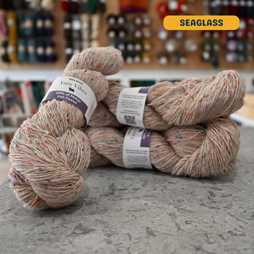100g beige with rainbow specks (seaglass) Irish wool merino sport yarn, 'Yarn Vibes Cliffs of Moher Irish Tweed' label visible, craft shop background (blurred)