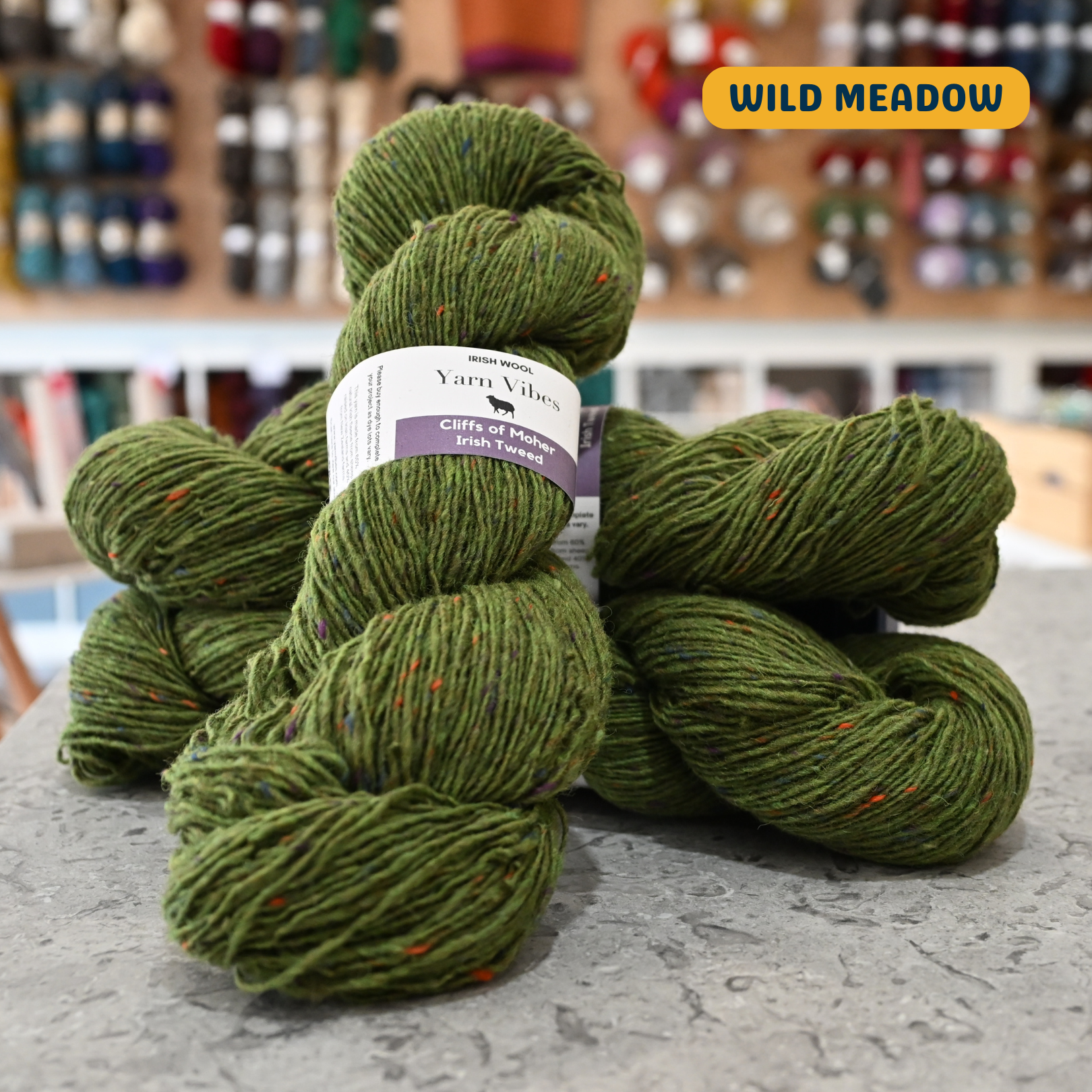 100g green  with rainbow specks (wild meadow) Irish wool merino sport yarn, 'Yarn Vibes Cliffs of Moher Irish Tweed' label visible, craft shop background (blurred)