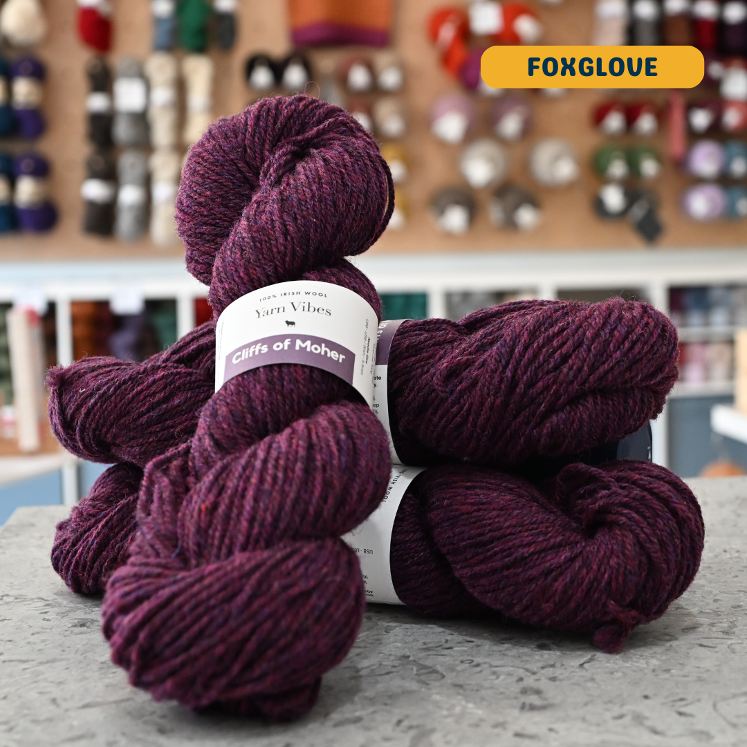 100g dark purple (foxglove) Irish wool  merino aran yarn, 'Yarn Vibes Cliffs of Moher' label visible, craft shop background (blurred)