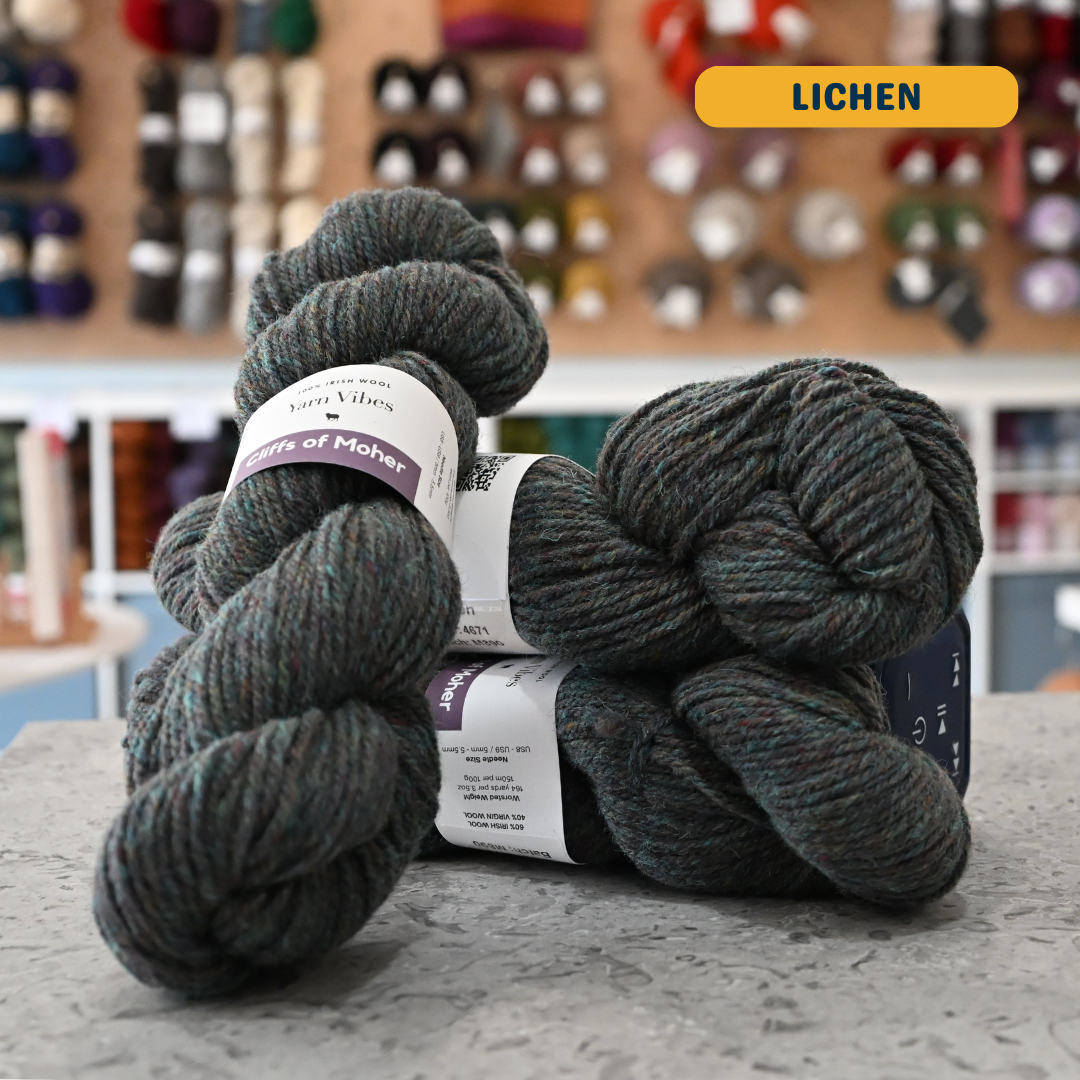 100g green-grey (lichen) Irish wool  merino aran yarn, 'Yarn Vibes Cliffs of Moher' label visible, craft shop background (blurred)
