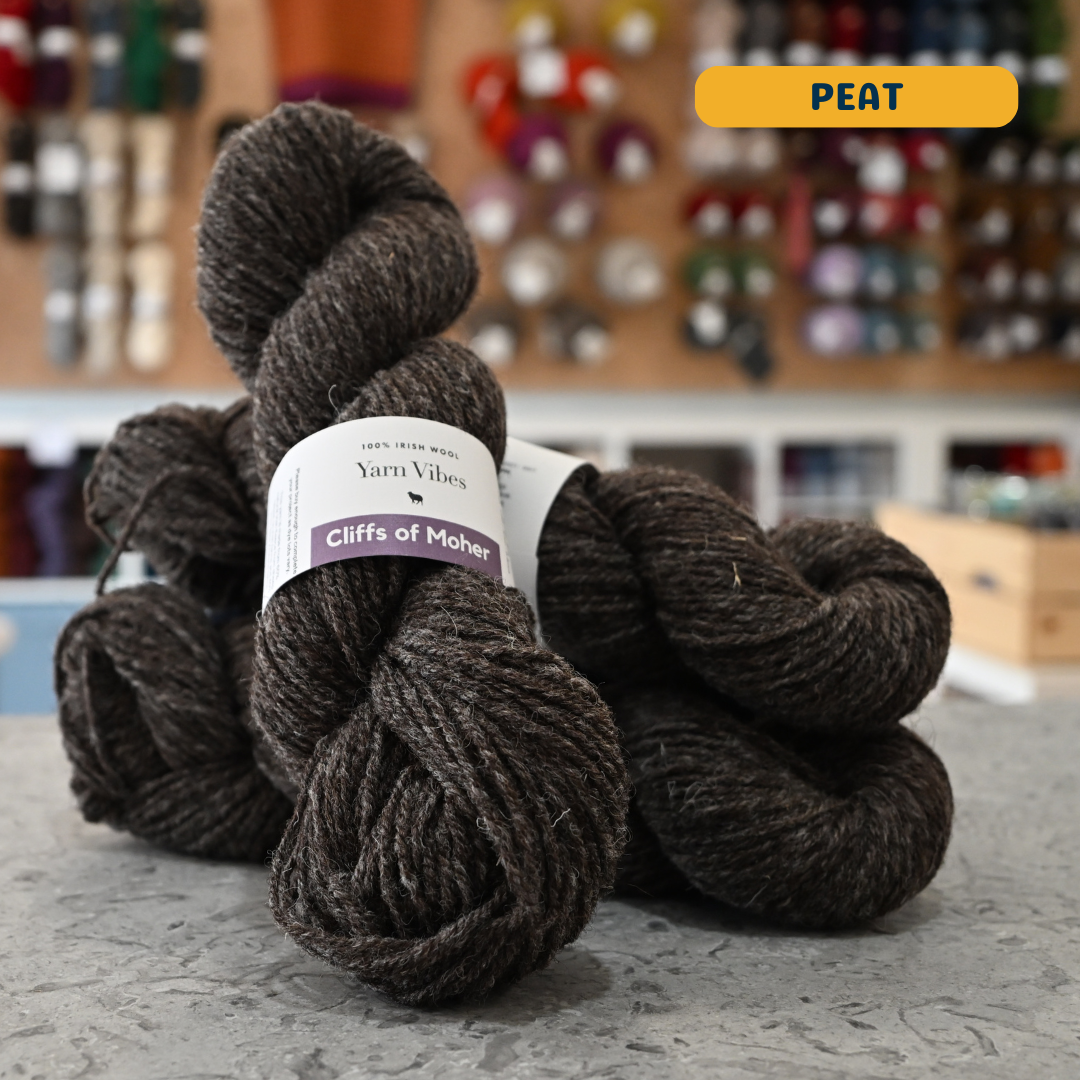 100g dark brown-grey (peat) Irish wool  merino aran yarn, 'Yarn Vibes Cliffs of Moher' label visible, craft shop background (blurred)
