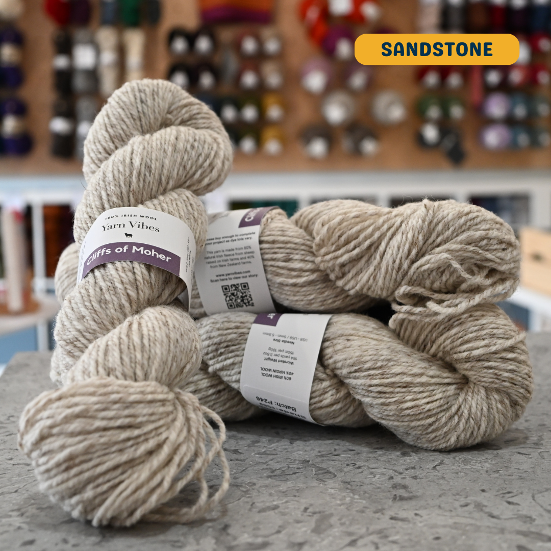 100g heathered beige (sandstone) Irish wool  merino aran yarn, 'Yarn Vibes Cliffs of Moher' label visible, craft shop background (blurred)